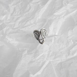 May include: A silver-colored ring featuring a heart-shaped design with angel wings. The ring has detailed feather textures and a small heart cutout at the center. The ring is set against a white, textured background.