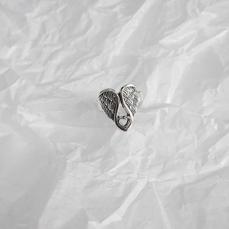 May include: A silver-colored ring featuring a heart-shaped design with angel wings. The ring has detailed feather textures and a small heart cutout at the center. The ring is set against a white, textured background.