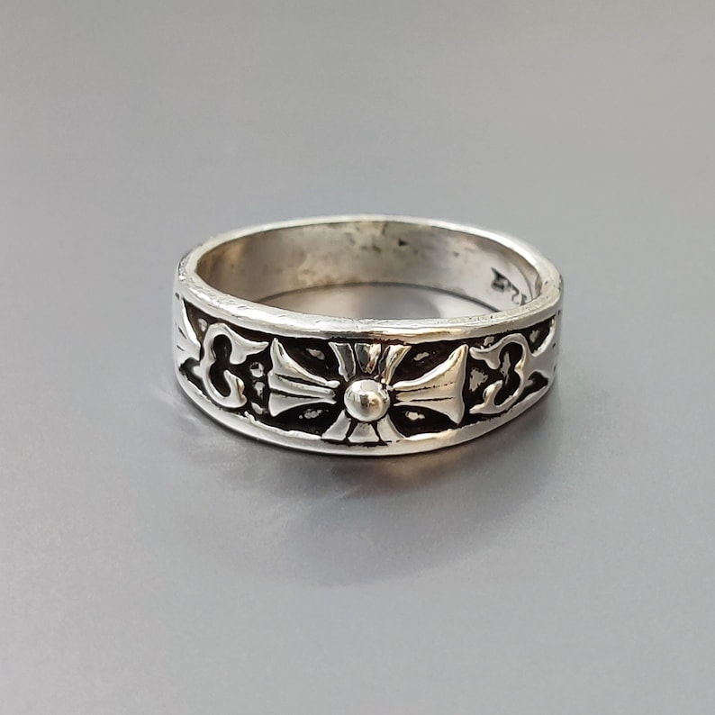 May include: A silver ring with a cross design. The cross is surrounded by intricate details and has a small bead in the center.