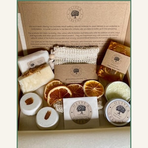 May include: A selection of natural toiletries in a gift box, featuring soaps, bath bombs, and body scrub. The box includes items with the "Olive & Greene" logo, dried orange slices, and a loofah. The products are vegan and cruelty-free.