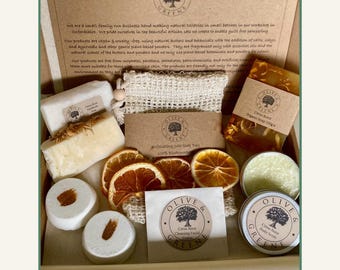 May include: A gift box filled with natural toiletries, including soaps, bath bombs, and body scrub. The box contains items with the "Olive & Greene" logo, dried orange slices, and a loofah. The products are vegan and cruelty-free.