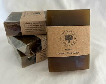 May include: A stack of four rectangular, dark green herbal organic soap bars. Each bar is wrapped in clear plastic and has a brown paper band with the text "OLIVE & GREENE Herbal Organic Soap 100g e". The soap bars are on a white surface.