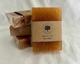 May include: A stack of four rectangular soaps, wrapped in clear plastic and brown paper. The front soap has a brown paper band with the text "OLIVE & GREENE" and "Down to Earth Soap 100g e". The soap is a light brown color.