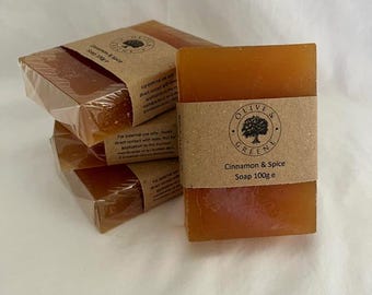 May include: Several bars of Cinnamon & Spice soap are displayed. The soap is a warm, amber color and wrapped in clear plastic. Each bar has a brown paper band with the Olive & Greene logo and product information. The label reads "Cinnamon & Spice Soap 100g e."