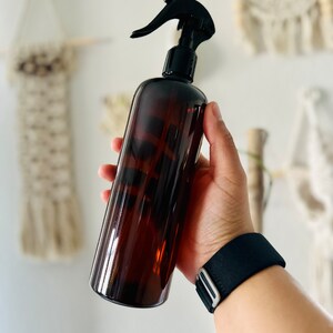 Puede incluir: Una botella de spray de vidrio marr&oacute;n con un pulverizador de gatillo negro. La botella est&aacute; siendo sostenida por una mano que lleva una pulsera negra.