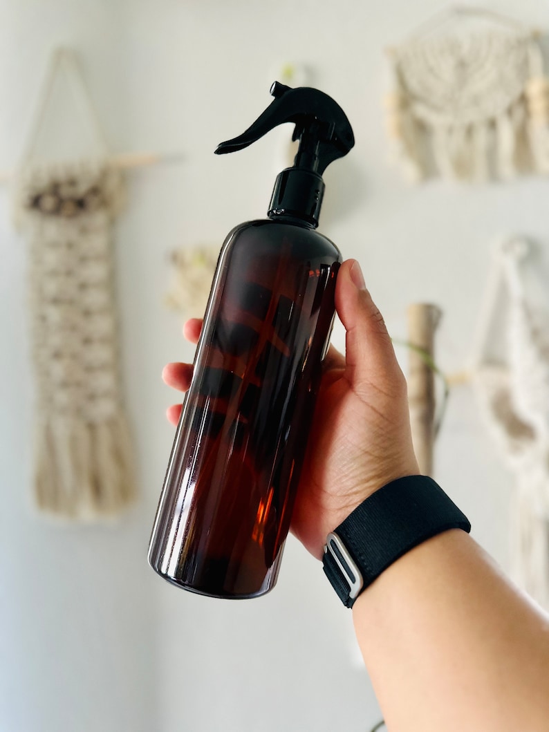 Puede incluir: Una botella de spray de vidrio marr&oacute;n con un pulverizador de gatillo negro. La botella est&aacute; siendo sostenida por una mano que lleva una pulsera negra.
