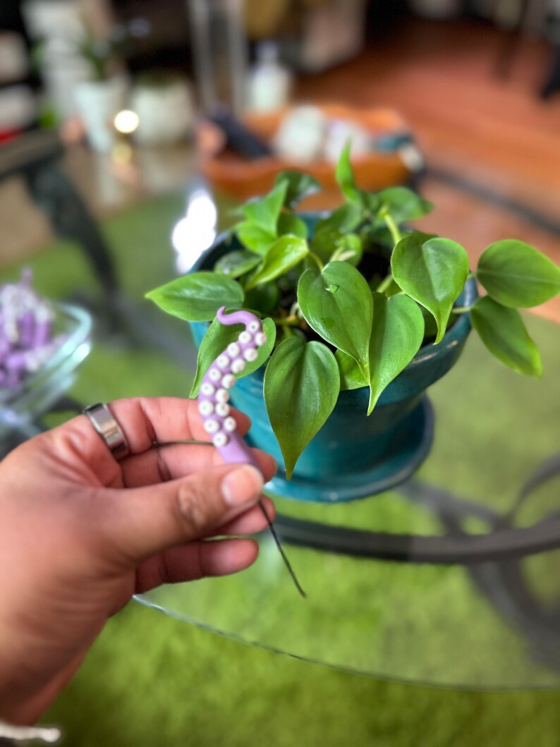 May include: A purple octopus-shaped pin with a black wire attached to the back. The pin is held in a hand, with a green potted plant in the background.