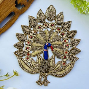 May include: Embroidered peacock applique with gold thread, blue body, and red accents. The peacock is surrounded by gold leaf-shaped details and small red flowers. This decorative applique is suitable for embellishing clothing or crafts.