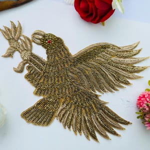 以下が含まれることがあります： 金色の刺繍が施されたワッペンで、複雑なビーズ細工とメタリックな糸のディテールが特徴です。翼を広げた横向きの鷲が枝をくわえており、目には小さな赤いビーズがあしらわれています。