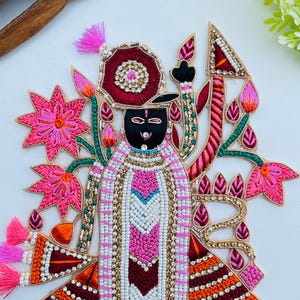 May include: Embroidered applique featuring a deity figure with a black face, adorned with pink, white, and gold embellishments. The design includes floral and geometric patterns, with pink tassels and a red and gold halo. The overall theme is religious.