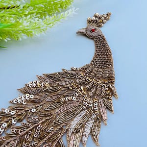 May include: A gold, brown, and white peacock applique with detailed beadwork and sequins. The peacock has a red gem eye and is set against a light blue backdrop. Green foliage is visible in the upper left corner, adding a natural element to the design.