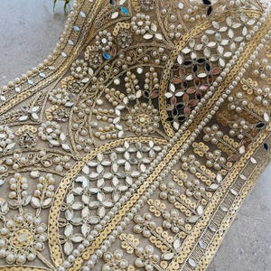 Può includere: Un bordino ricamato in oro e bianco con perline e specchietti. Il bordino presenta un motivo ripetitivo di disegni floreali e forme geometriche.