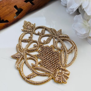 May include: Gold-colored embroidered applique with a floral design. The applique features intricate detailing, including woven and looped elements, and is likely used for embellishing garments or crafts. The design includes leaves and a central fan-like shape.