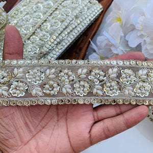 May include: A close-up of a delicate, off-white lace trim with gold accents and pearl-like beads. The trim features a floral design and is perfect for adding a touch of elegance to any project.