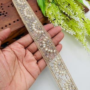 May include: A decorative trim featuring a cream-colored mesh base, adorned with faux pearls and sparkling rhinestones. The trim is bordered by a gold-colored, intricate design. This trim is suitable for embellishing garments or craft projects.