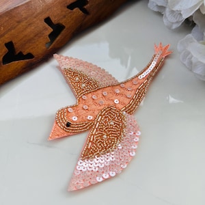 May include: A peach-coloured, embroidered bird appliqué with beaded and sequined details. The bird's wings are spread in flight, with a mix of gold and pink sequins. The bird's body is outlined with gold beads and has a small black eye.