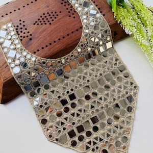 Puede incluir: Un collar decorativo beige y plateado con un patrón geométrico de espejos. El collar está hecho de tela beige y está adornado con espejos de varias formas, incluyendo cuadrados, círculos y triángulos. El collar está diseñado para ser usado alrededor del cuello.