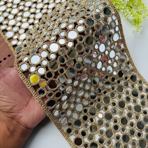 May include: Gold-colored trim with embedded circular mirrors. The trim has a woven design with small, reflective mirrors evenly spaced throughout. The edges are finished with a delicate gold thread, creating a decorative border. This trim is suitable for embellishing garments or crafts.