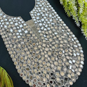 Puede incluir: Una pieza decorativa plateada para prendas de vestir, muy adornada con pequeñas formas reflectantes en forma de espejo, de diversas formas geométricas. La pieza tiene una base redondeada y un escote redondo, con un respaldo de malla.