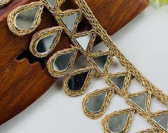 May include: Close-up of gold-colored trim with mirrored accents. The trim features a repeating pattern of triangular and teardrop-shaped mirrored pieces, each outlined with gold thread. The trim is likely used for embellishing clothing or craft projects.