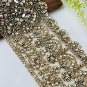 May include: Close-up of a decorative trim featuring intricate beadwork. The trim has a cream-colored base with gold-colored metallic thread and is embellished with pearls, clear and gold-colored rhinestones, and mirrored accents. The design is ornate and elegant.