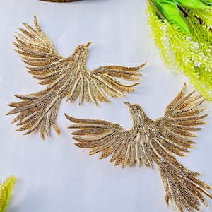 May include: Two gold-colored bird embellishments with outstretched wings and long tail feathers. The birds are intricately detailed with a beaded texture, suggesting a handcrafted design. The background is white, with green and white floral accents.