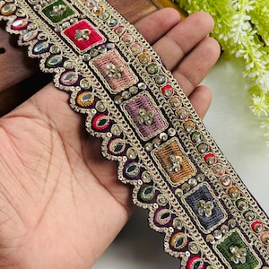 Puede incluir: Un adorno decorativo con una base morada oscura, que presenta cuadrados bordados en varios colores como rojo, verde y azul, adornados con lentejuelas y pequeños espejos. El adorno tiene un borde festoneado y detalles de hilo dorado, para ropa o manualidades.