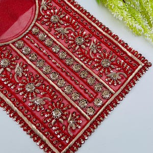 May include: A red, embroidered textile with gold-colored floral and geometric patterns. The fabric is adorned with sequins and intricate threadwork, creating a decorative design. The textile has a rounded neckline.