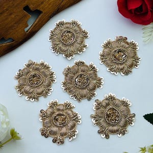Peut inclure: Six écussons brodés circulaires de couleur dorée avec un motif floral. Chaque écusson est réalisé avec du fil métallique, du fil torsadé et de petites paillettes disposées en pétales autour d'un centre texturé en relief à bord festonné.