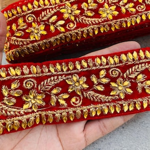 May include: Close-up of red fabric trim with gold floral embroidery and sparkling embellishments. The trim features detailed designs of flowers and leaves, with a border of small, golden gems. The fabric has a velvet-like texture.