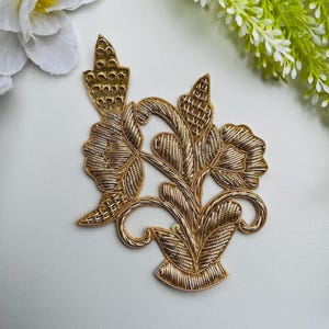 Può includere: Applicazione floreale dorata con dettagli intricati. Il design presenta fiori, foglie e steli stilizzati, il tutto realizzato con una trama tessuta o ricamata. L'applicazione è probabilmente destinata ad abbellire abiti o progetti artigianali.