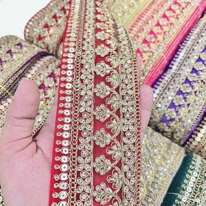 May include: A red fabric trim featuring intricate gold metallic embroidery in a floral pattern and a border of small, circular gold sequins. The decorative ribbon is made of a sheer mesh material and is displayed alongside other colorful rolls.
