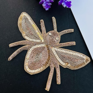 May include: A decorative, beaded insect applique in shades of gold and brown. The wings and body are intricately detailed with sequins and beads. The applique is set against a black background with purple flowers.