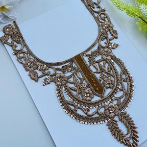 以下が含まれることがあります： 金色の刺繍が施された、装飾的なネックラインアップリケ。 花と葉の模様が特徴で、スパンコールで装飾されています。 アップリケは、中央に茶色のパネルと、装飾的で細長い下部要素を備えています。 白い背景に配置されています。