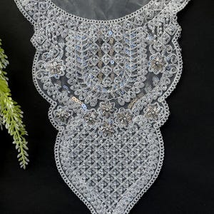 Puede incluir: Aplicación de encaje blanco con cuentas y lentejuelas. El diseño presenta un borde festoneado, patrones florales y geométricos, y una forma de lágrima en la parte inferior. La aplicación es probablemente para embellecer ropa o accesorios.