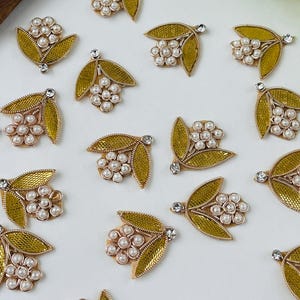May include: Close-up of several decorative floral embellishments. Each piece features a gold-colored leaf design with a cluster of white pearls and a small, clear rhinestone. The embellishments are scattered on a white surface.