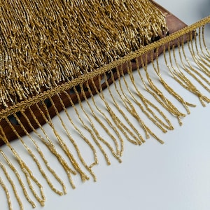 May include: Close-up of gold beaded fringe trim. The trim features long strands of gold beads attached to a woven gold band. The fringe is densely packed, creating a luxurious and textured appearance. The trim is likely used for embellishing clothing or home decor items.