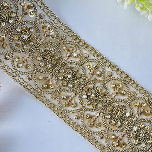 Peut inclure: Garniture brodée dorée avec un motif floral et géométrique. La garniture présente des paillettes et des perles dorées, créant un aspect luxueux et orné. La garniture est sur un support en résille crème.