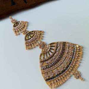 May include: A gold-coloured beaded embellishment with detailed patterns. The design includes a fan-shaped element with rows of beads and sequins, and floral details. This embellishment is likely for decorative use on clothing or accessories.