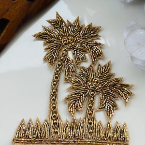 May include: Gold beaded palm tree applique with detailed leaves and trunks, and a beaded grass base. The applique is likely for embellishing clothing or accessories, adding a touch of tropical flair.
