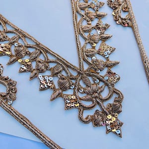 May include: Gold embroidered trim with floral and leaf patterns. The trim is decorated with sequins and has a braided edge. The detailed design and metallic finish suggest it could be used for embellishing clothing or crafts.