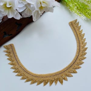 May include: Gold-colored beaded applique with a leaf-like design. The applique is in a curved shape, with a detailed pattern of small beads and a brown border. The background is white with white flowers and green foliage.