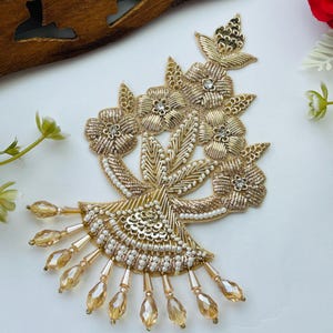 May include: A gold embroidered floral applique featuring metallic thread, sequins, and small white beads. The design includes five flowers with central clear crystals and a fan-shaped base adorned with dangling, faceted amber-colored glass beads.