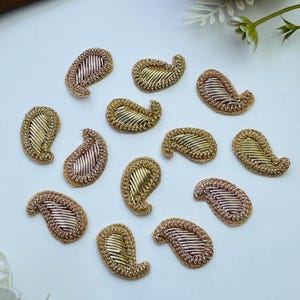 May include: A collection of twelve small paisley-shaped embroidered patches. Each patch features metallic gold and silver thread work with a raised, textured border and diagonal stitching patterns, arranged in a scattered formation on a white surface.