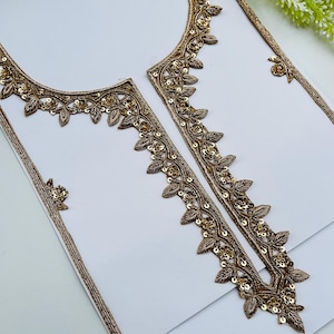 Może przedstawiać: Zbliżenie białej tkaniny z misternym złotym haftem. Haft ma kwiatowy i liściasty wzór z cekinami. Projekt jest prawdopodobnie przeznaczony na odzież, z widocznym dekoltem i obramowaniem.