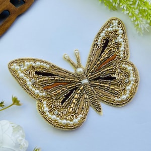 May include: A gold-colored, beaded butterfly applique with pearl and rhinestone accents. The butterfly has intricate detailing, with brown and orange accents on the wings. The wings are outlined with small pearls. This decorative applique is suitable for clothing or craft projects.
