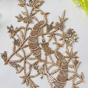 May include: Embroidered peacock design with gold and silver sequins, beads, and thread. The design features two peacocks perched on branches with leaves. The birds have red accents and are surrounded by floral elements. This decorative piece is suitable for embellishing garments or crafts.
