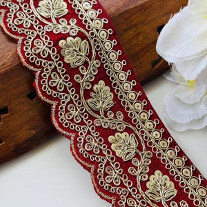 May include: Close-up of a decorative red ribbon with gold embroidery. The ribbon features a scalloped edge and intricate floral and circular patterns. The design is detailed and elegant, suitable for embellishing crafts or garments.