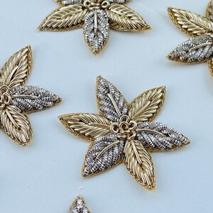 May include: Close-up of several gold and silver floral embellishments. Each flower has six petals, with gold-colored wire outlining the petals and silver-colored detailing in the center. The flowers are arranged on a light background.