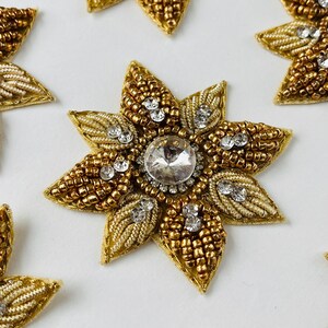 Peut inclure: Gros plan de plusieurs ornements floraux en perles dorées. Chaque fleur a huit pétales, un grand strass transparent au centre et de plus petits strass sur les pétales. Les pétales sont faits de perles dorées et de fil d'or.